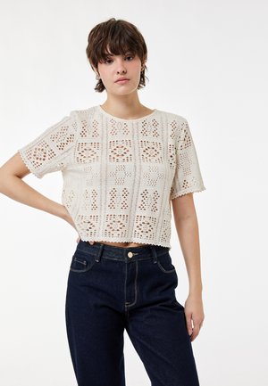 Jeune personne aux cheveux bruns courts portant un haut blanc en crochet à manches courtes et un jean taille haute bleu foncé, debout avec la main sur la hanche.