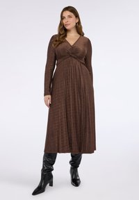 Brun klänning med lång ärm, V-ringning och knytdetalj i midjan, med en flödande kjol och texturerat tyg. Matchad med svarta knähöga stövlar.