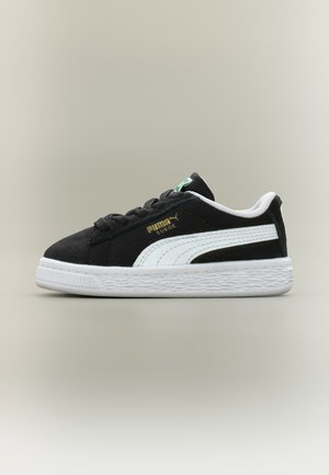 Czarny but sportowy Puma Suede z białym paskiem Puma, czarnymi sznurowadłami i fakturowaną białą podeszwą na jednolitym jasnym tle.