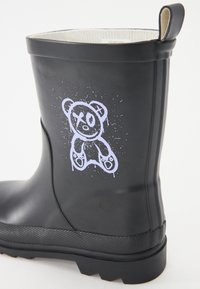 Čierne gumové dažďové čižmy s textúrovanou podrážkou. Obsahujú grafiku bieleho medveďa a rozliate farby na boku. Hladký povrch a zaoblený vrch.