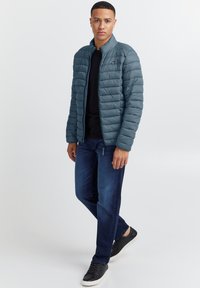 Polstret, glidelåsjakke i dyp smaragdblå med horisontale sømmer; brukt over en svart skjorte og mørke jeans. Har en ståkrage og minimalistisk design.