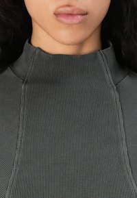 Hnedý ribovaný top s vysokým golierom, ktorý má textúrovanú tkaninu a priliehavý strih. Švy pozdĺž strán posilňujú štruktúru.
