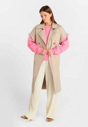 Femme vêtue d'un trench-coat sans manches beige sur un sweat-shirt rose, d'un pantalon large texturé crème et de talons ouverts marron, debout sur fond blanc.
