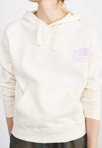 Personne portant un sweat à capuche blanc cassé avec poche avant et logo "The North Face Patron Plasticfree Peaks" en violet clair sur la poitrine.