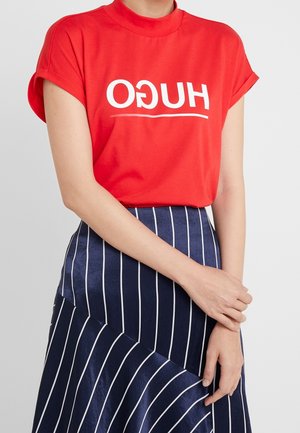Persoon draagt een rood t-shirt met omgekeerde witte "HUGO" tekst en een marineblauwe rok met witte verticale strepen, handen samengevouwen voor zich.