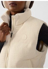 Gilet in pile color crema con colletto alto, chiusura con cerniera e tessuto testurizzato. Presenta una toppa con logo e uno strato inferiore in maglia scura.