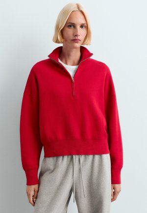 Femme aux cheveux blonds courts portant un pull rouge à fermeture éclair quart et un pantalon gris clair à cordon sur un fond clair uni.