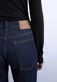 Personne portant un jean en denim bleu foncé avec des coutures jaunes et un haut à manches longues côtelé noir, montrant la poche arrière et l'étiquette de marque en cuir.