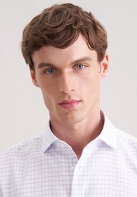 Weißes Hemd mit Knöpfen, das ein hellblaues Karomuster, einen taillierten Kragen und eine markante Textur aufweist, getragen von einem Modell mit kurzen, welligen Haaren.