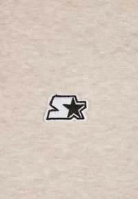 Tela beige con un pequeño logo bordado en blanco y negro, que representa una letra 'S' estilizada junto a una estrella.