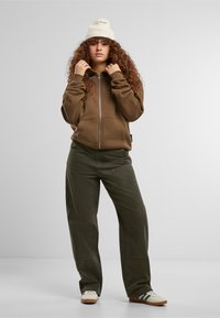Brauner Zip-Hoodie mit einer strukturierten crema-farbigen Strickmütze, lockeren olivgrünen Hosen und weißen Schuhen mit grünen und dunkelblauen Akzenten.
