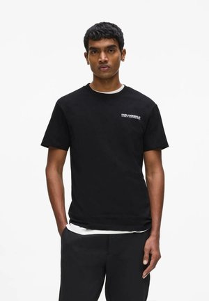 Mladý muž stojaci čelom vpred, oblečený v čierne tričko Karl Lagerfeld a čierne nohavice, ruky v vreckách, neutrálny výraz.