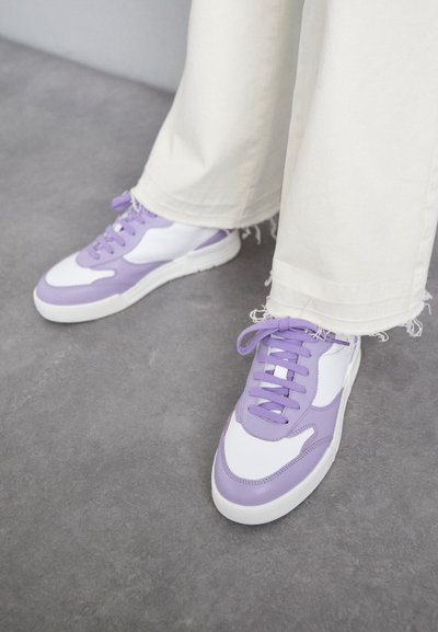 Lila och vita sneakers med en slät läderöverdel, texturerade accents och platta snören. Matchas med trasiga vita vida byxor.