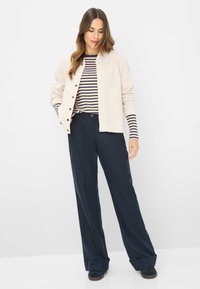 Beige knoopjack over een marineblauw en wit gestreept hemd met lange mouwen, gecombineerd met wijde marineblauwe broek en donkere schoenen.
