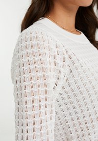 Pull en tricot blanc avec un motif texturé en maille ouverte. Présente un col rond et des poignets côtelés. Le tissu semble léger et respirant.