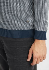 Virkat tröja i marinblått och ljusblått med ett texturerat mönster, ribbade ärmslut och en liten logodetalj nära fållen. Bärs med svarta byxor.