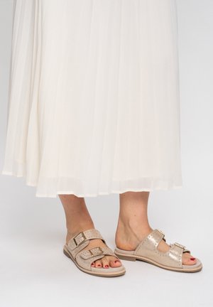 Pieds nus avec du vernis à ongles rouge portant des sandales plates beiges à double boucle, partiellement couverts par une jupe blanche fluide.