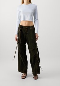 Kvinde iført hvid langærmet cropped top, mørkegrønne vide bukser med cargo-lommer og stropper samt sorte højhælede sandaler med stropper.