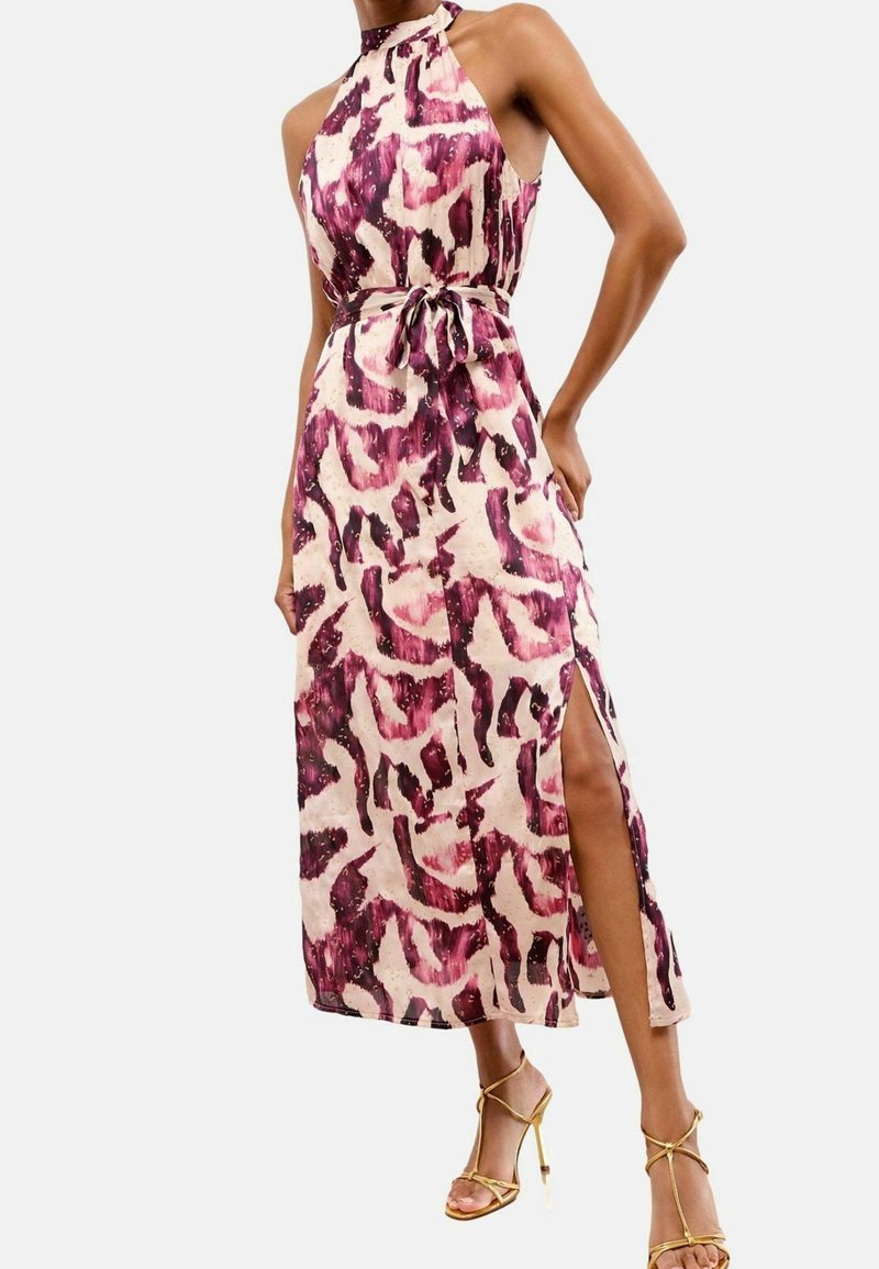 Femme porte une robe à motifs roses et violets, sans manches, à col licou, avec une ceinture à la taille et une fente latérale, associée à des sandales dorées à talons hauts.