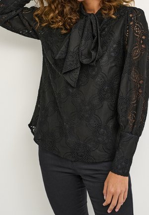 Donna che indossa una blusa nera di pizzo con motivi floreali e maniche a palloncino, abbinata a pantaloni neri, mentre posa con un braccio sollevato.