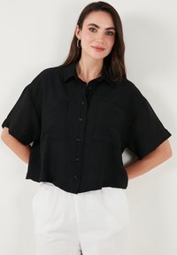 Camicia nera a maniche corte con bottoni, realizzata in tessuto testurizzato, con due tasche sul petto e un design accorciato. Indossata con pantaloni chiari.