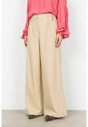 Ženska, oblečena v široke bež hlače, rožnato dolg rokav bluzo in črno-bele čevlje z zebra vzorcem, stoji na betonski tlakovani površini.