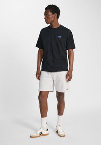 Zwarte katoenen t-shirt met een blauw logo op de linkerborst, gecombineerd met lichtgrijze shorts. Witte sneakers met zwarte accenten en dikke witte sokken.
