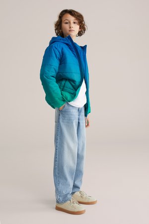 Otrok z kodrastimi lasmi, oblečen v modro-zelen puhasti plašč, belo majico, svetlo modre kavbojke in bež superge, stoji pred bež ozadjem.