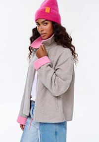 Šedá fleecová bunda s růžovými akcenty na límci a manžetách, s zipovým zapínáním a bočními kapsami. Doplněná růžovou čepicí.