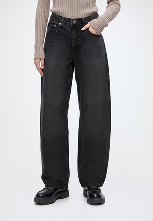 Personne portant un jean noir taille haute à jambes larges, des chaussures noires épaisses et un pull beige côtelé à manches longues, main dans la poche.