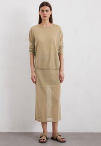 Haut beige à manches longues avec une coupe décontractée et un col arrondi, associé à une jupe longue semi-transparente assortie et des sandales plates.