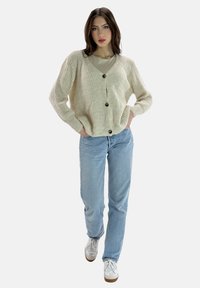 Cremfarbener Strickcardigan mit V-Ausschnitt, großen braunen Knöpfen, lockerer Passform und geripptem Textur, kombiniert mit hellblauen Jeans.