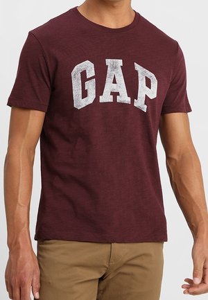 Mann trägt ein kastanienbraunes T-Shirt mit kurzen Ärmeln und verblasstem weißen "GAP"-Logo sowie khakifarbene Hosen vor einem einfarbigen Hintergrund.