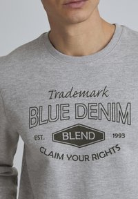 Sudadera gris con cuello redondo, que presenta un logo impreso en negro que dice "Trademark Blue Denim Blend Claim Your Rights" en forma de diamante.
