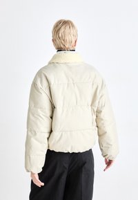 Veste rembourrée beige avec un col en peluche, dotée de sections matelassées, de manches longues et d'une coupe décontractée. La vue de dos montre un design simple sans embellissements.