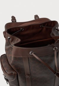 Zaino in pelle marrone con una superficie strutturata a ciottoli e rifiniture lisce. Presenta una chiusura con cordoncino e cuciture più chiare in contrasto.