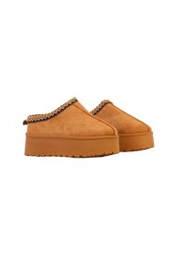 Lysebrune mokkasiner i ruskind med tyk, flad sål. Overdelen har en dekorativ vævet kant langs kanten, som giver en struktureret detalje.