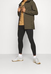 Olivgrüne wasserdichte Jacke mit Kapuze, kombiniert mit schwarzen Leggings und hellgrauen Sportschuhen. Darunter trägt sie ein bedrucktes beiges T-Shirt.