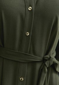 Olijfgroene jurk met een gladde textuur, knoopsluiting en een gestrikte riem. Voorzien van glanzende zilveren knopen en een gestructureerde kraag.
