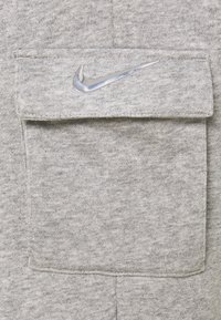 Grå hoodie-tyg med en mjuk textur. Har en framficka med en sydd klaff och en glänsande silverfärgad Nike-logotyp på klaffen.
