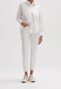 Vit skjorta med knappar och en bröstficka, kombinerad med vita smala byxor och ljusa sneakers, alla med en slät textur.