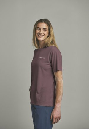 Junge Frau mit hellbraunen Haaren, die lächelt, trägt ein lila T-Shirt und blaue Hose, steht vor einem schlichten grauen Hintergrund.