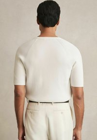 Top cu mâneci scurte, tricotat cu nervuri, în nuanță albă, cu un design cu mâneci raglan și un decolteu curat, asociat cu pantaloni de culoare deschisă.