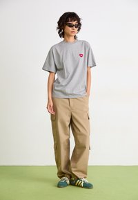 Person iført grå T-skjorte med rødt hjerte-logo, beige cargo bukser, teal sneakers og svarte solbriller, står på grønt gulv mot hvit vegg.