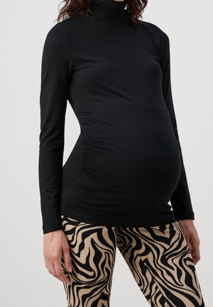 Zwangere persoon die een nauwsluitende zwarte coltrui en beige-zwart zebraprint broek draagt, zichtbaar van schouders tot dijen.