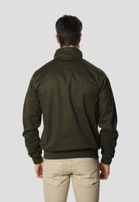 Zielona kurtka bomberka z prążkowanym kołnierzem i mankietami, o luźnym kroju i naszywką na ramieniu. Nosi się ją na jasnobeżowych spodniach.