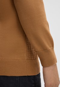Brauner Langarmpullover mit Bündchen und Saum sowie strukturiertem Seitenpaneel, getragen über dunklen Hosen, zeigt einen Teil des Arms und der Hand einer Person.