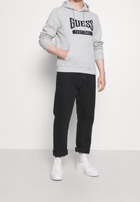 Sudadera gris con texto negro "GUESS", bolsillo delantero y capucha con cordón, combinada con jeans negros ajustados y zapatillas blancas.