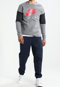 Harmaa huppari mustilla hihoilla, jossa on suuri punainen Nike-logo ja "AIR"-teksti. Yhdistettynä tummansinisiin housuihin ja valkoisiin lenkkareihin.