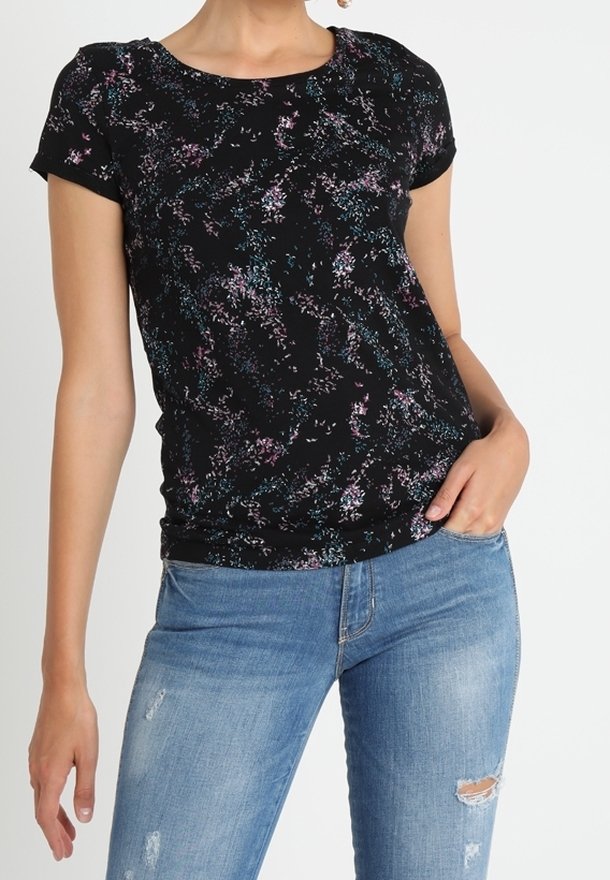 Femme portant un haut noir à manches courtes avec un motif abstrait multicolore et un jean skinny bleu clair déchiré.